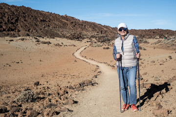 Active cheerful senior woman with backpack walking outdoors with help of poles enjoying nature, freedom and free time. Arid terrain at the base of Teide volcano on the island of Tenerife