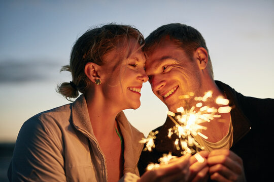 Couple, happy and beach fireworks with sunset for anniversary celebration, outdoor date and love. Smile, man and woman with sparks by ocean for romantic vacation, honeymoon and healthy relationship