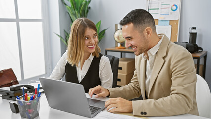 A smiling woman and man, colleagues in conversation while looking at a laptop in a modern office setting.