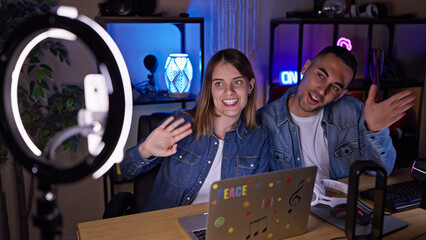 A man and woman engage as content creators in a well-equipped, modern home office with a ring light, waving at their online audience.
