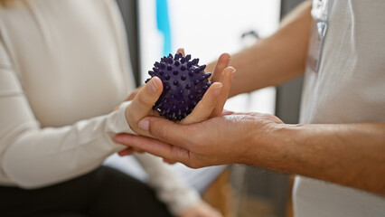 A woman therapist assists a man patient with a spiky purple massage ball in a bright rehabilitation center.