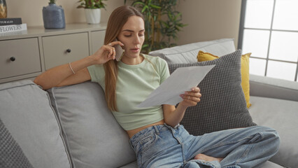 Beautiful young woman with blonde hair talking on her phone and reading a document while sitting on a sofa in a cozy living room.