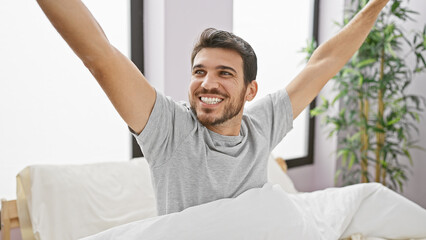 A cheerful young hispanic man waking up and stretching with a broad smile in a sunlit modern bedroom