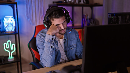 A pensive young man wearing headphones sits in a dark gaming room illuminated by neon lights. © Krakenimages.com