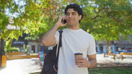 A young hispanic man enjoys a conversation on his mobile phone while holding a coffee cup in an urban park.