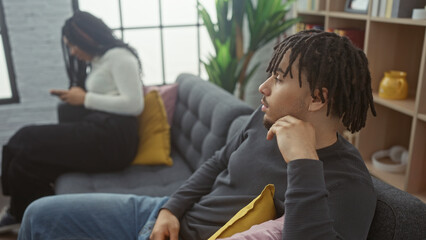 A concerned man and a woman using a phone sit together in a well-lit living room, depicting modern relationship dynamics indoors.