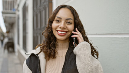 A young smiling hispanic woman talking on a mobile phone while standing on an urban street.