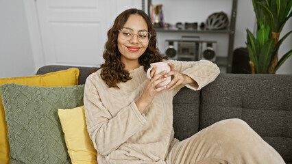 A smiling woman in glasses holds a pink mug, sitting on a couch with yellow pillows in a cozy...