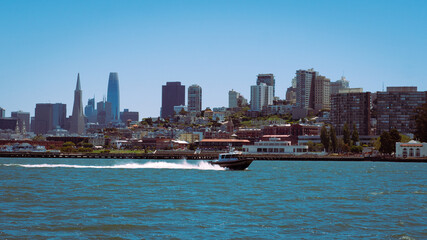 Obraz premium San Francisco skyline, including the Transamerica Pyramid and Salesforce Tower, with a boat speeding across the water in the foreground. The scene is set under a clear blue sky.