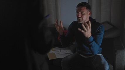 A worried man gestures during an intense interrogation by an unseen policewoman in a dimly lit...