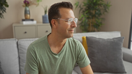 Hispanic man sitting in a cozy living room with green shirt and glasses, looking thoughtful