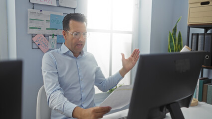 Man wearing glasses discussing a document with expressive gestures in a bright office environment, surrounded by charts and notes on the wall, at a work desk with a computer monitor.
