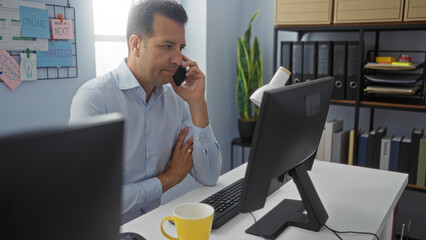 Hispanic man talking on a mobile phone in an office, looking at a computer monitor with a serious expression, surrounded by organized folders and decor in a well-lit environment.