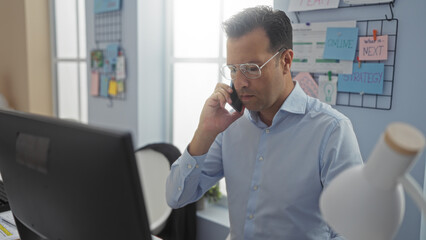 Obraz premium Middle-aged hispanic man working in an office while talking on the phone, surrounded by strategy notes and a computer, depicting a focused and professional workplace setting.
