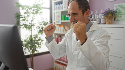 A happy hispanic middle-aged male pharmacist celebrates at his computer in a well-stocked pharmacy interior.