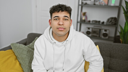 A handsome young man wearing a white hoodie comfortably seated on a grey couch indoors, exuding a casual, relaxed atmosphere.