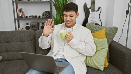 A relaxed young man waving during a video call at home, with laptop and coffee mug on the couch.