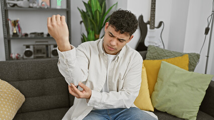 A young, bearded man feeling elbow pain while sitting on a couch indoors.