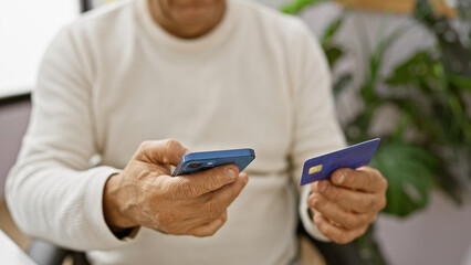 A middle-aged hispanic man using a smartphone and holding a credit card indoors, likely in an office setting.