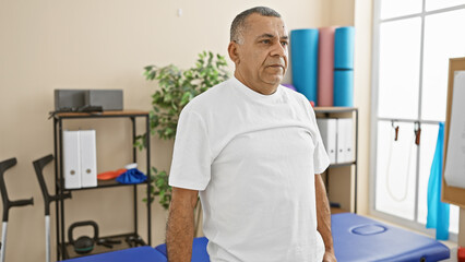 Mature hispanic man standing contemplatively in a bright rehabilitation clinic interior.