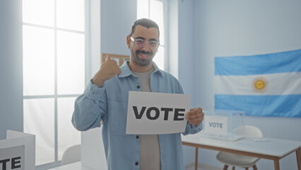 Young hispanic man voting in an argentinian electoral room with a flag and banner in the background, holding a vote sign and showing a thumbs-up gesture indoors.