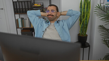 Young man with moustache wearing glasses relaxing in office chair in a modern workplace with plants and shelves in the background