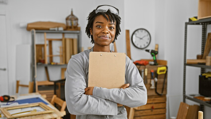 Confident young woman with dreadlocks wearing safety glasses in a woodworking workshop, holding a clipboard.