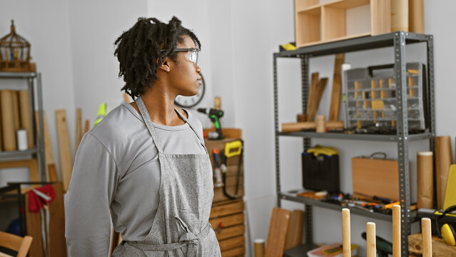 A young african american woman with dreadlocks wearing a smock stands contemplatively in a well-organized carpentry workshop.