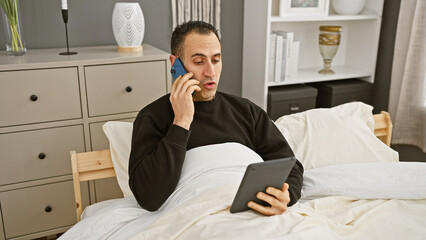Hispanic man multitasking with phone and tablet while sitting in a well-adorned bedroom.