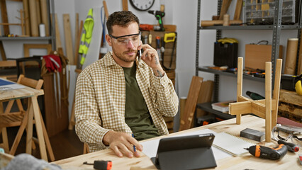 A young hispanic man with a beard talks on the phone in a carpentry workshop, surrounded by tools and wood.