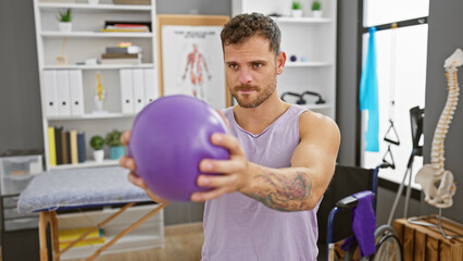 Handsome hispanic man with beard doing physical therapy exercises with a purple ball inside a clinic.