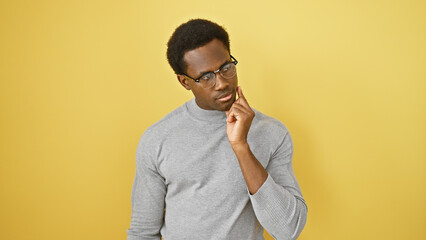 A thoughtful young african american man wearing glasses, isolated against a yellow background,...