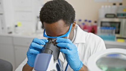 African american male scientist using microscope in laboratory setting, wearing blue gloves and white coat.