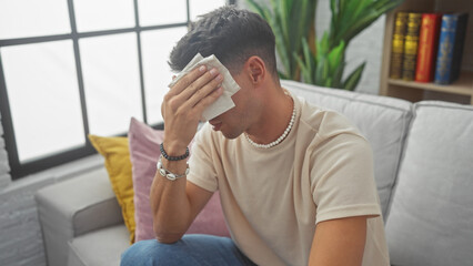 A distressed young hispanic man sitting in a modern living room, wiping his forehead in contemplation or illness.