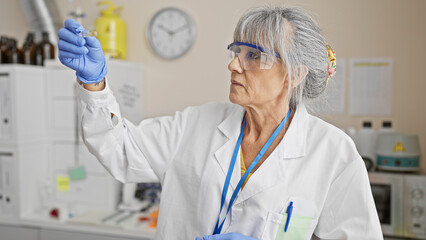 A mature woman scientist analyzes a sample in a laboratory, wearing safety glasses and gloves.