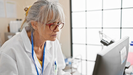 Mature woman in glasses focuses on a computer screen within the bright, white space of a modern hospital lab