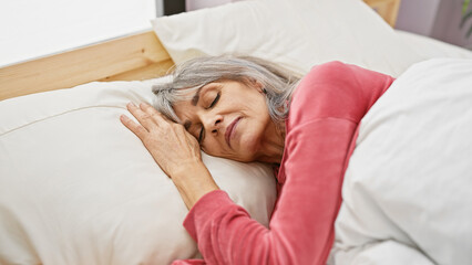 A serene middle-aged woman with grey hair sleeps peacefully in a cozy bedroom setting, indicating rest and comfort.