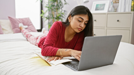 Naklejka premium Young, south asian woman in casual attire working on a laptop while lying on a bed in a well-lit bedroom.