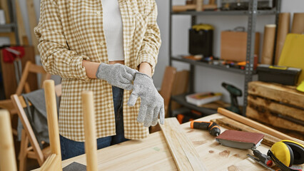 A woman wearing safety gloves preparing to work on a carpentry project in a workshop.