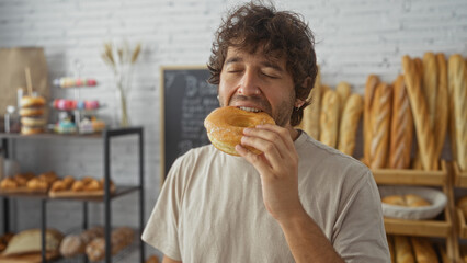 Handsome young man enjoying a fresh donut in a cozy bakery filled with various pastries and bread.