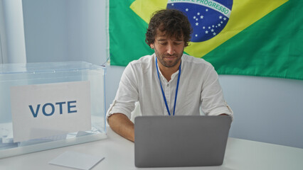 Young man using laptop at electoral college table with voting box, brazilian flag, and indoors setting.