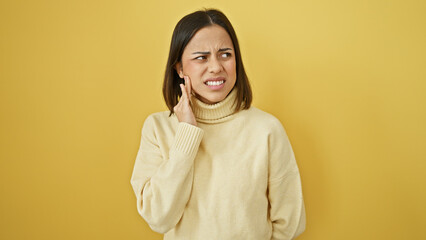 Young hispanic woman with a pained expression touching her cheek isolated against a yellow wall...