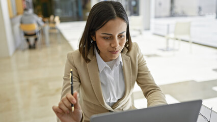 A focused hispanic woman in business attire works attentively on a laptop in a modern office environment.