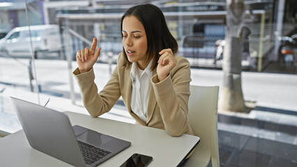 Hispanic woman in a beige blazer gestures while working on a laptop at a modern office with glass...