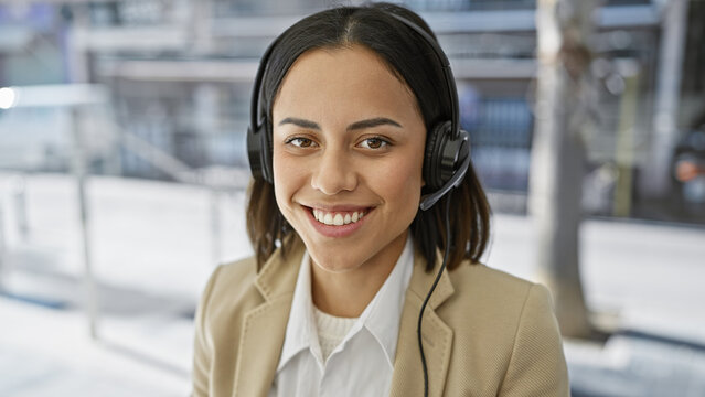 Smiling young hispanic woman wearing a headset in a professional setting with a blurred background.