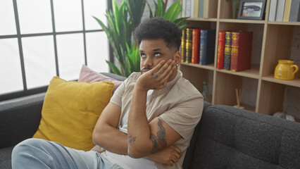 Thoughtful african american man seated indoors on a gray sofa, surrounded by home decor and plants