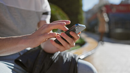 Obraz premium A young hispanic man casually using a smartphone outdoors on a sunny day, portraying connectivity in urban settings.