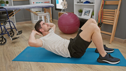 Hispanic man doing sit-ups on a blue mat inside a rehab clinic, with therapy equipment around.