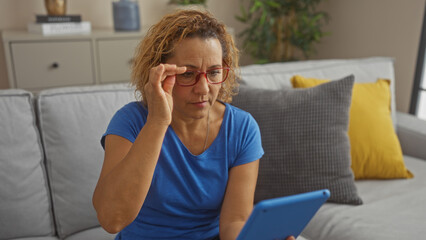 Middle-aged hispanic woman with curly hair wearing glasses and blue shirt uses a tablet in her living room.