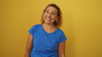 Obraz premium Beautiful mature hispanic woman with curly hair smiling and looking up, isolated over a yellow background.